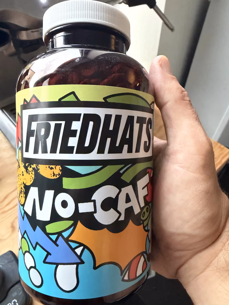 A dark glass jar of Friedhats No-Caf coffee beans, held in one hand in front of an espresso machine.