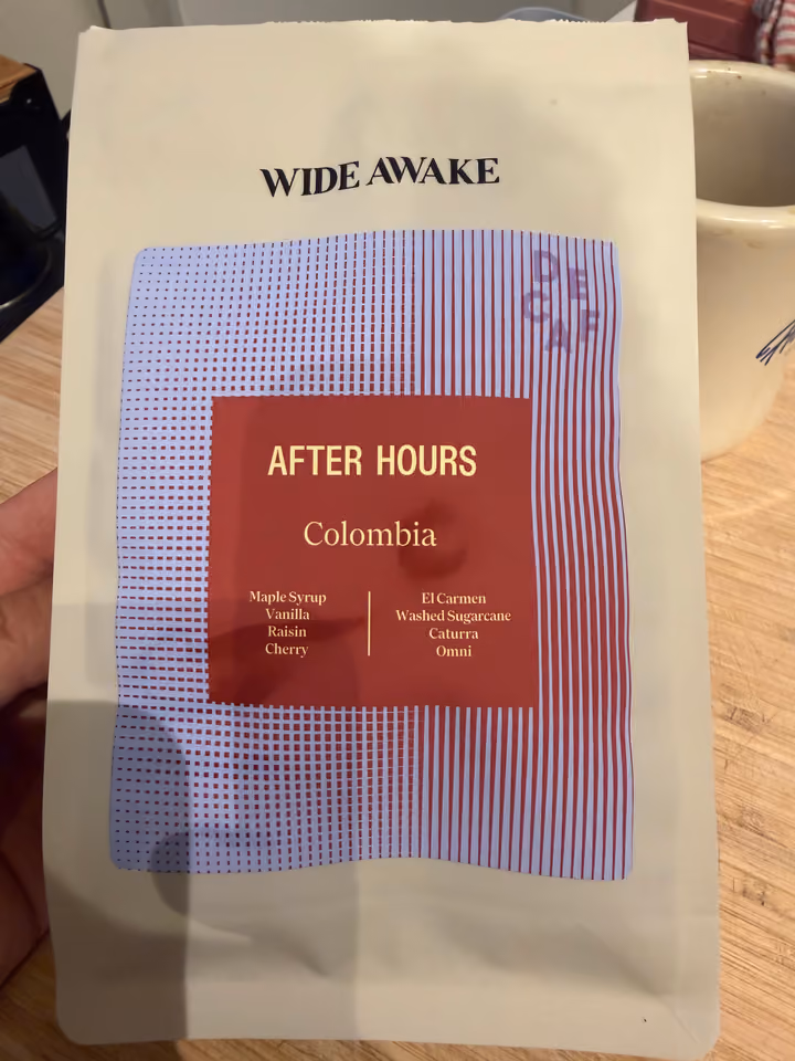 A cream Wide Awake bag of After Hours Colombia decaf held in one hand over a wooden worktop, with a mug behind it.